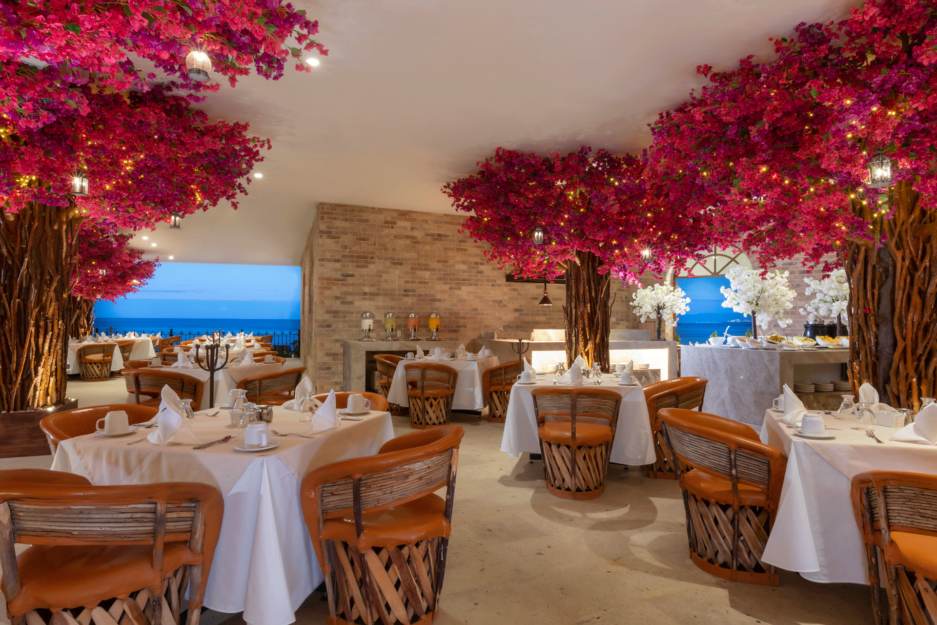 Elegant restaurant with white-clothed tables, orange chairs, and decorative pink bougainvillea trees overlooking ocean at Playa Los Arcos Puerto Vallarta 2023