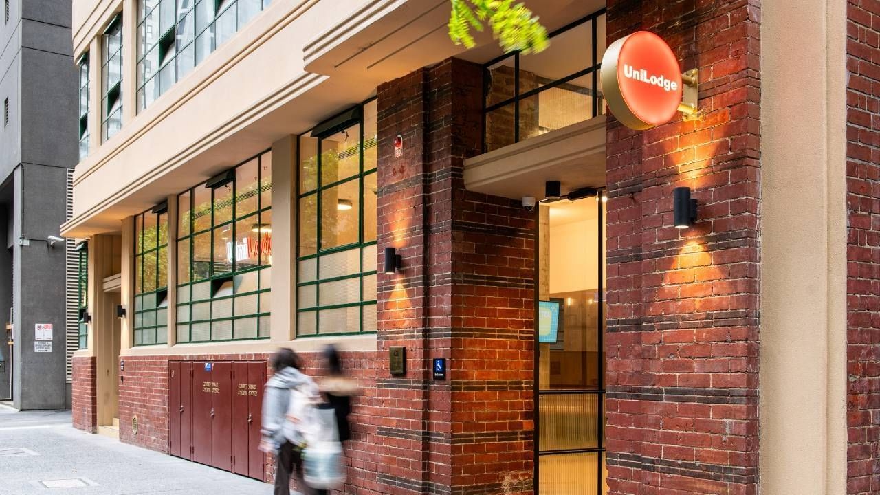 Two people walking past a brick building entrance with a UniLodge sign and green windows.