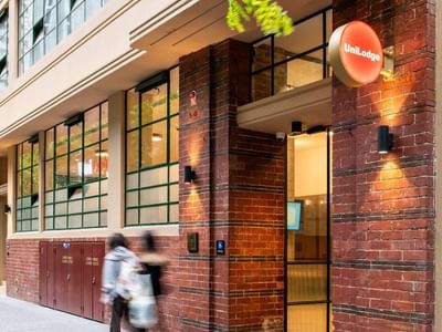 Two people walking past a brick building entrance with a UniLodge sign and green windows.