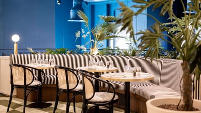 A restaurant with a blue wall, a striped bench, tables with chairs, and plants.