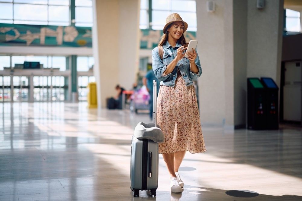 A woman in a hat, skirt, and denim jacket scrolls on her phone in an airport beside her carry-on suitcase.