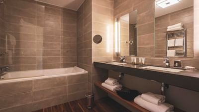 Modern bathroom with large bathtub, dual vanity, tiled walls, wooden floor, and multiple towels.