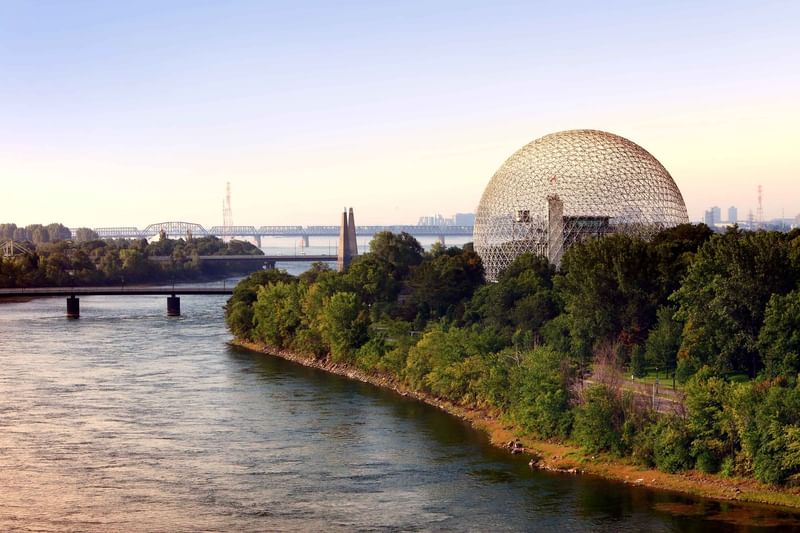 Geodesic dome structure surrounded by green trees along a river with bridges and city skyline at Warwick Le Crystal Montreal