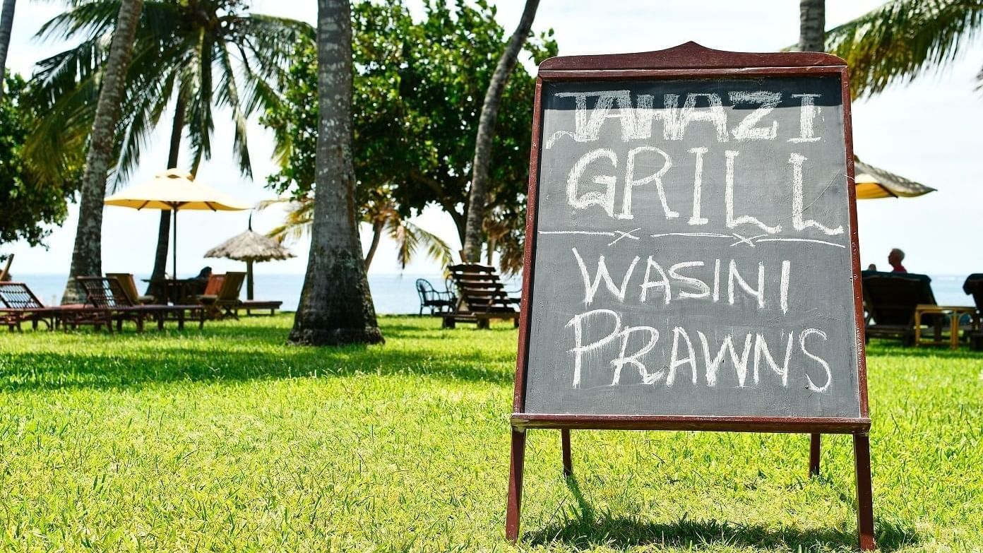 Chalkboard sign for Jahazi Grill on green lawn with palm trees and ocean views in the background at Serena Hotels & Resorts