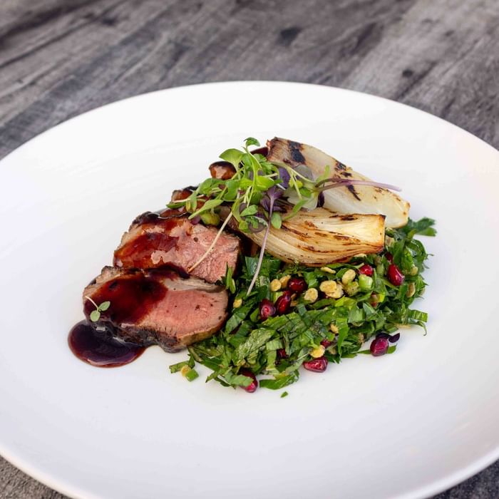 Plate with sliced meat, red sauce, green salad with pomegranate seeds, and grilled fennel on wooden table.