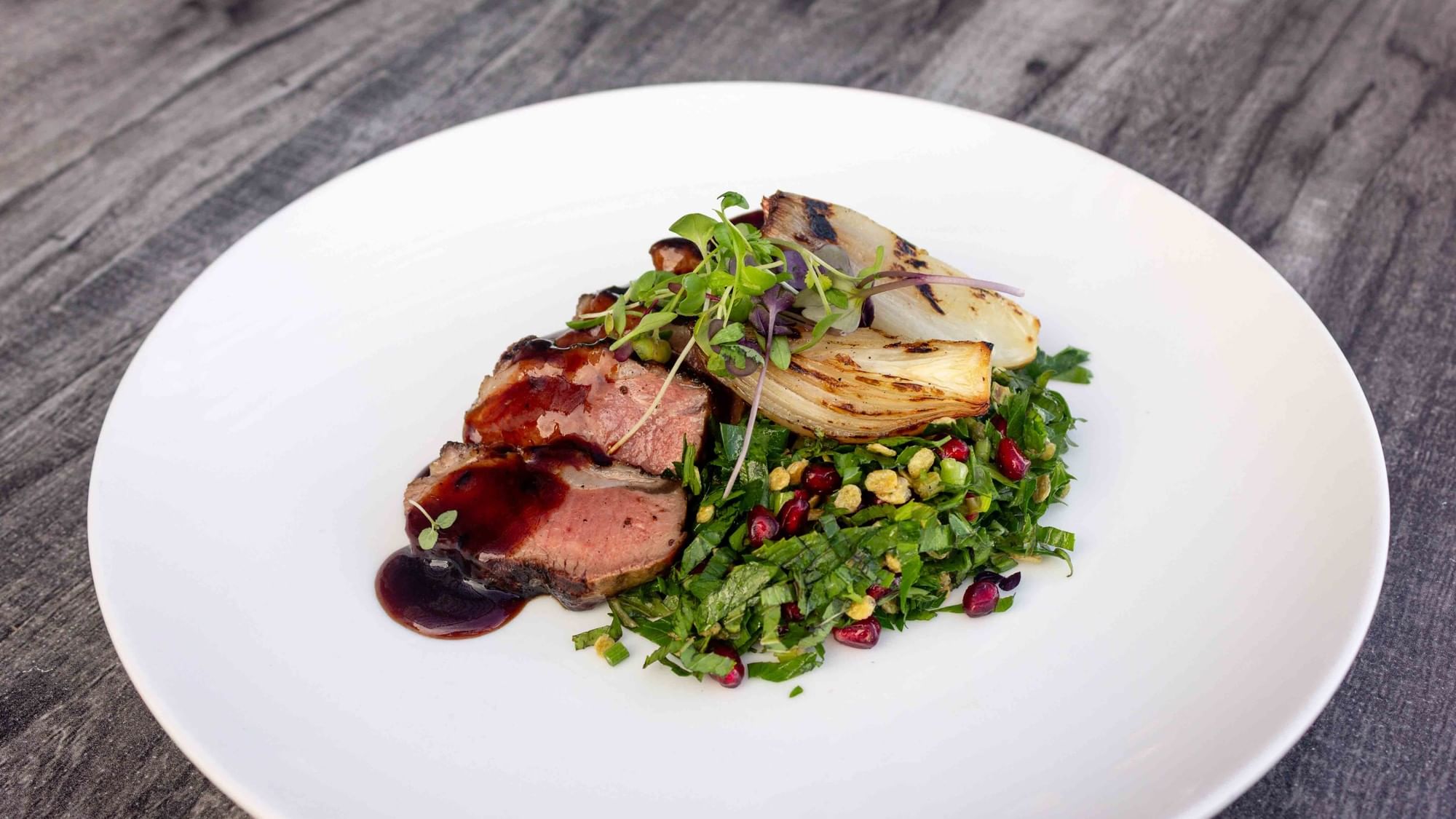 Plate with sliced meat, red sauce, green salad with pomegranate seeds, and grilled fennel on wooden table.