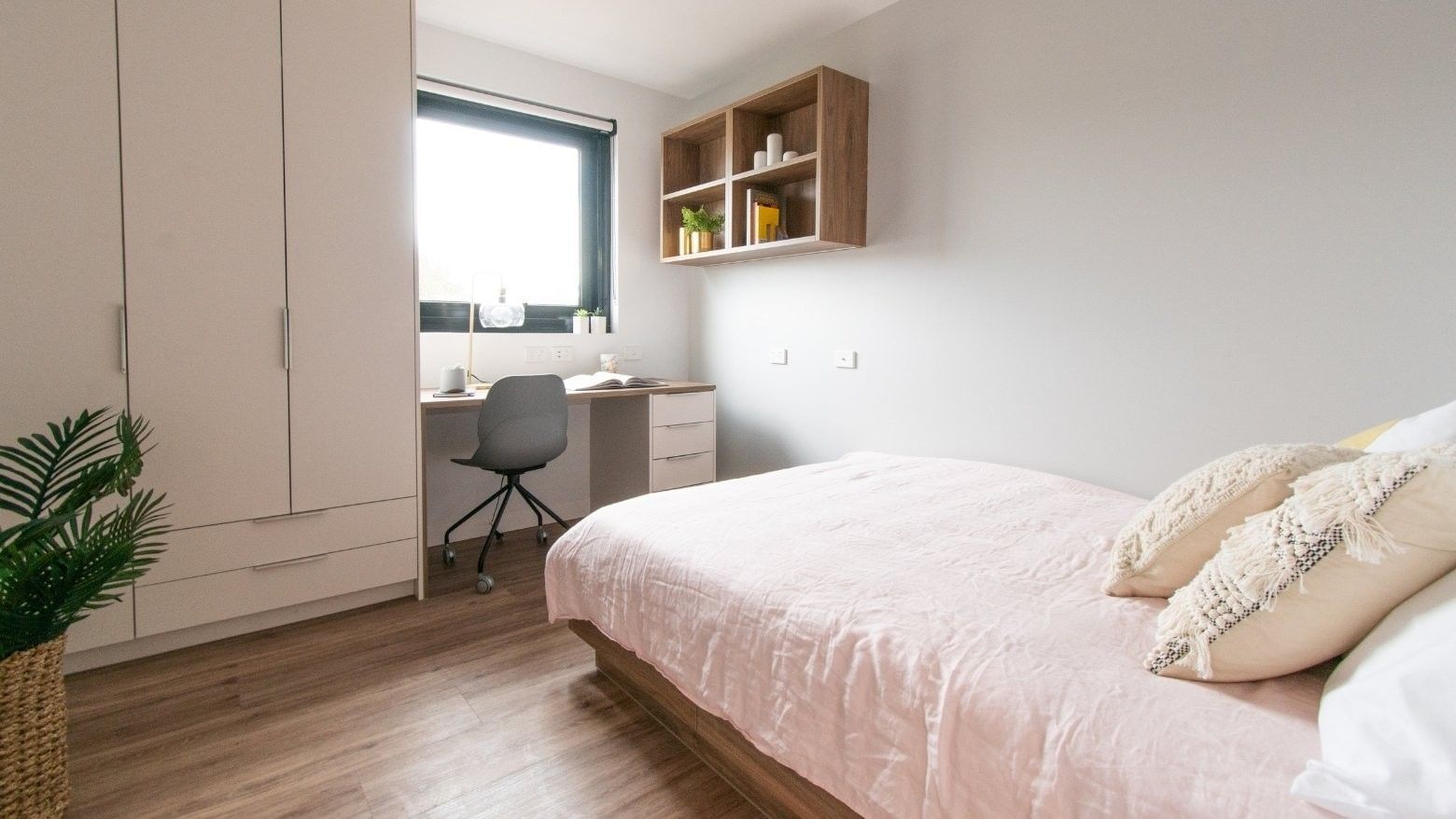 Modern bedroom with a pink bed, desk, chair, and cabinets, featuring a plant and window.