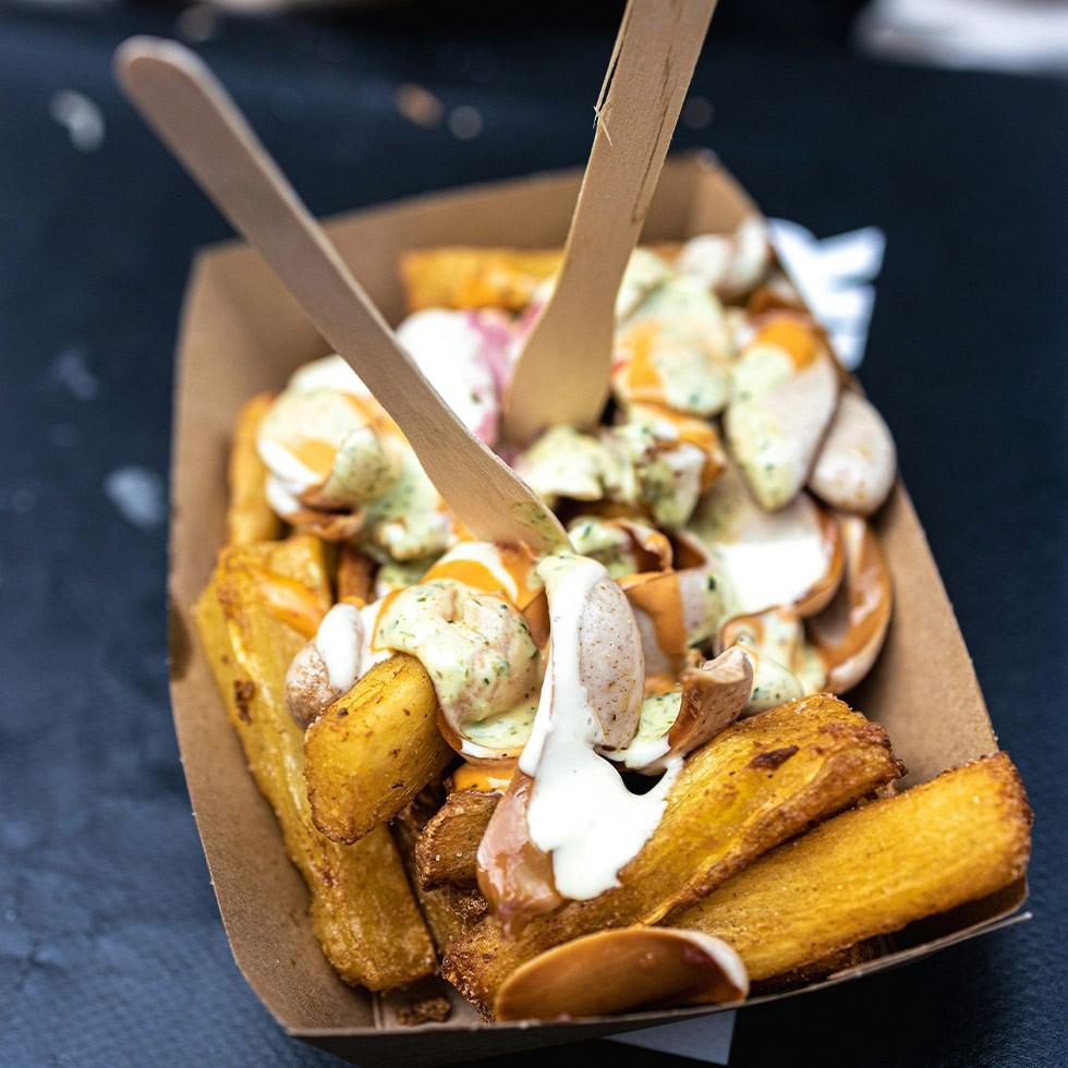 A carton of fried potatoes with toppings and two wooden spoons at European Street Food Festival.