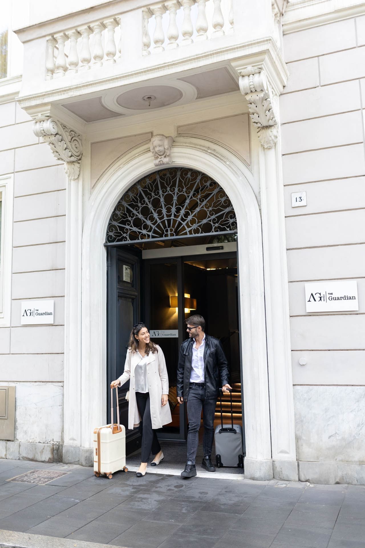 A couple exits The Guardian Hotel with ornate architecture and a sign, carrying small bags