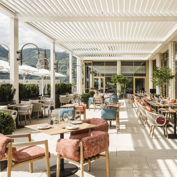 Eine Terrasse mit Tischen und Stühlen unter einem weißen, gebogenen Pergoladach und mit Blick auf die Berge.