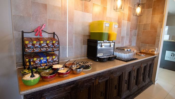 Cereal boxes and juice dispensers beside topping bowls on a wooden buffet counter in Palmira Cafeteria at Araiza Calafia