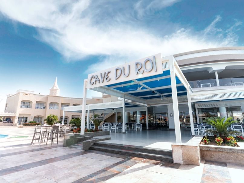 Exterior of Cave Du Roi restaurant with outdoor seating area and pool in the background.