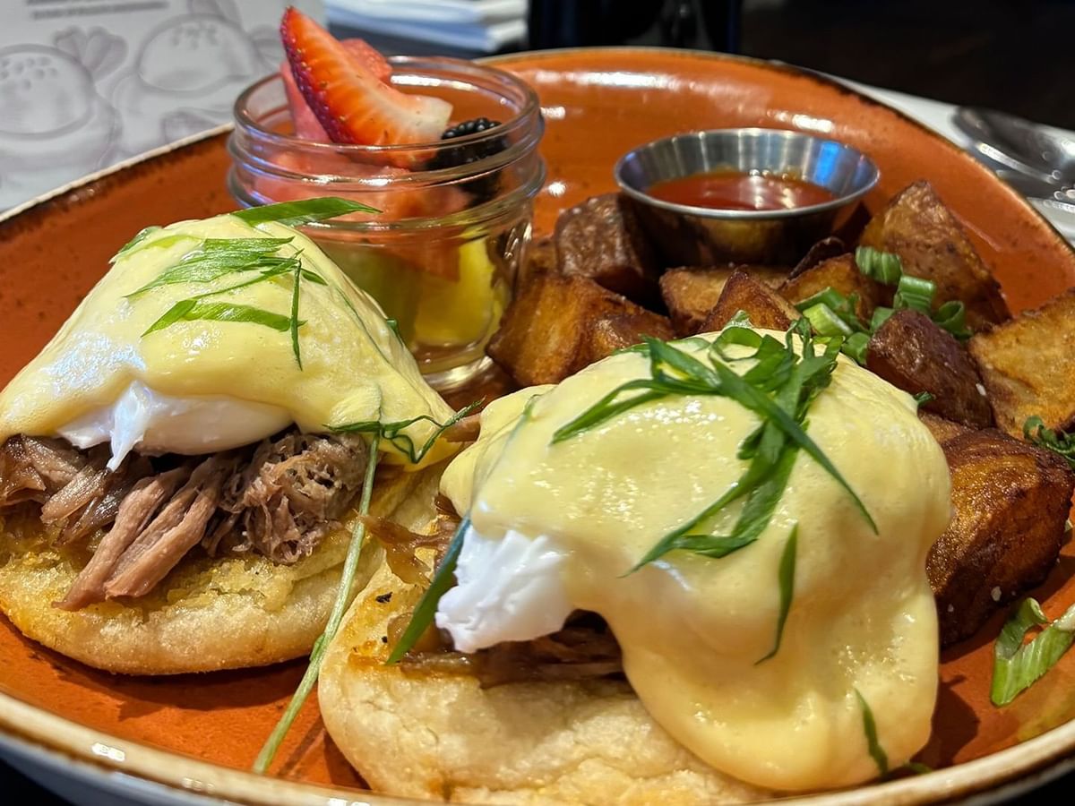 A plate with two egg benedict sandwiches, a side of potatoes, and a fruit cup.