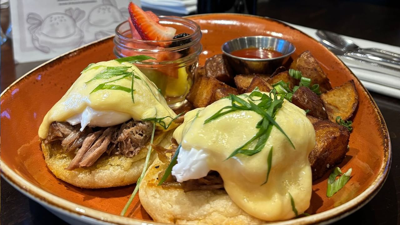A plate with two egg benedict sandwiches, a side of potatoes, and a fruit cup.