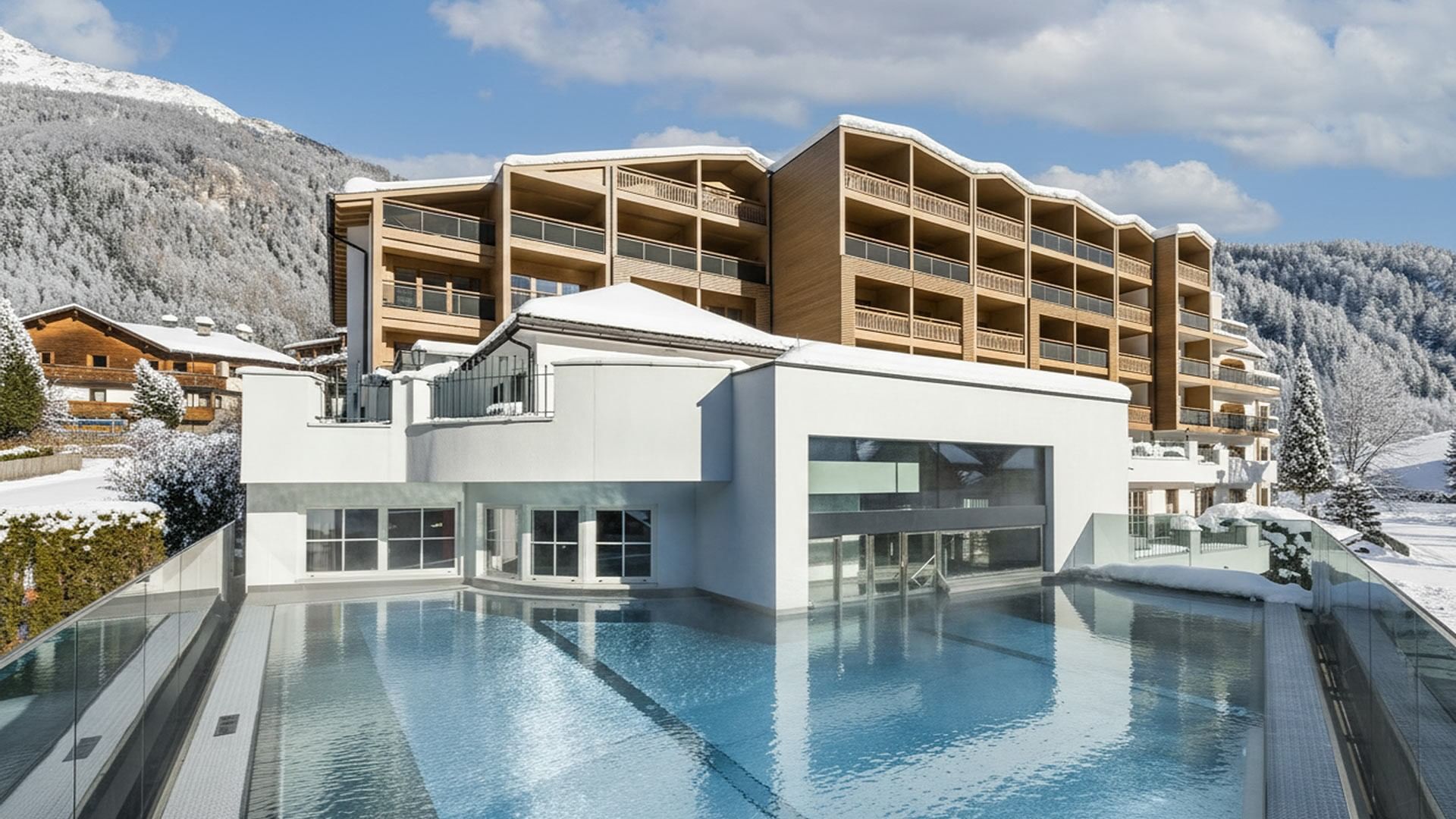 Piscina esterna in un resort di montagna con edifici moderni e neve sulle colline circostanti.