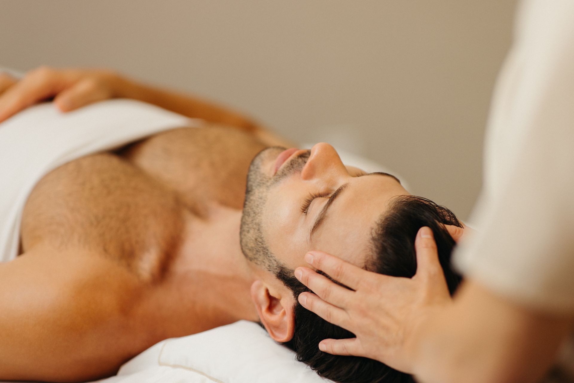 Man with closed eyes lying on a massage table receives head massage from a therapist.