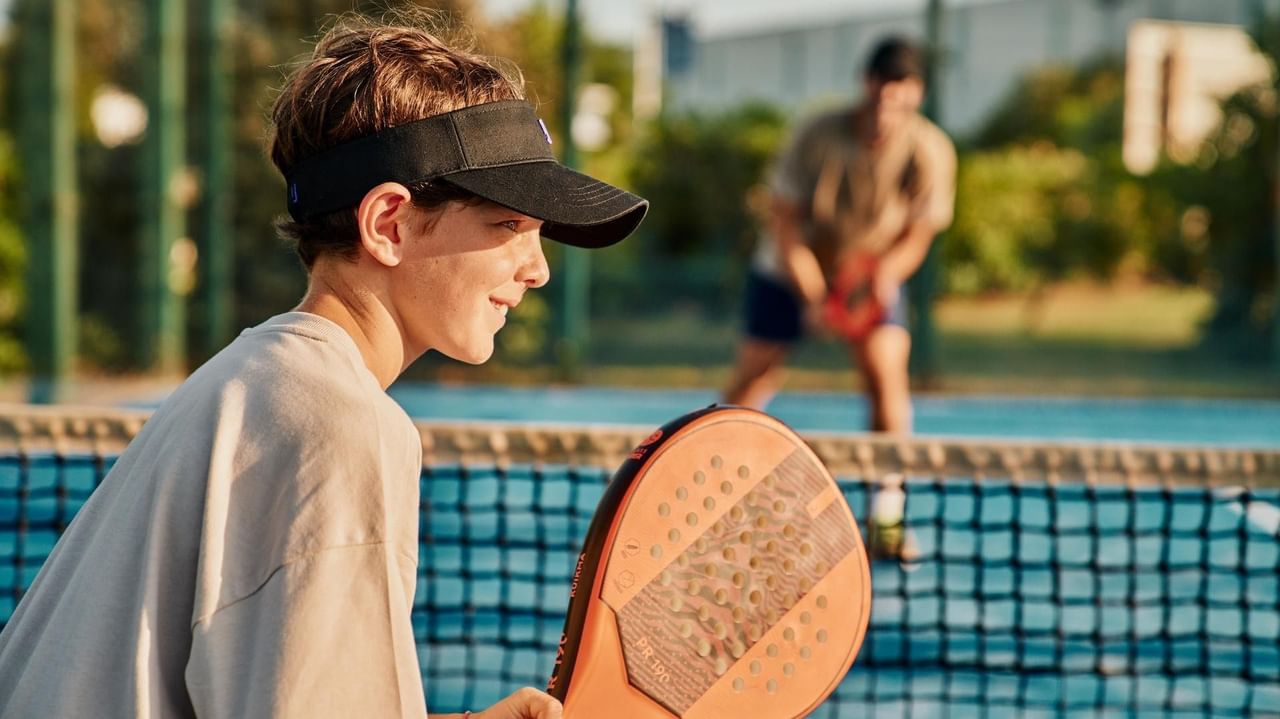 Junge Person mit Visier und Schläger auf einem Padelplatz, ein anderer Spieler im Hintergrund.