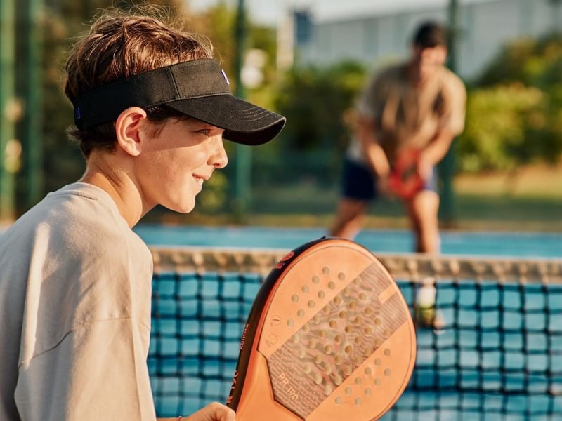 Junge Person mit Visier und Schläger auf einem Padelplatz, ein anderer Spieler im Hintergrund.