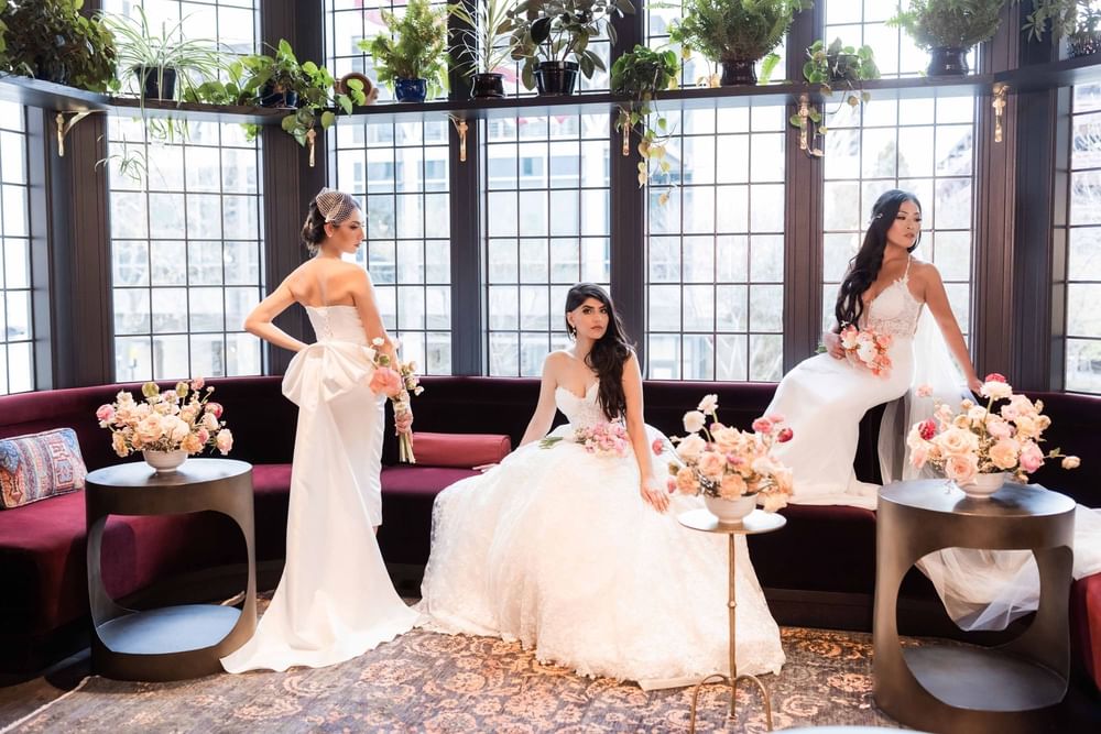 Three stunning brides in elegant gowns posing gracefully amid beautiful floral arrangements at El Prado Hotel