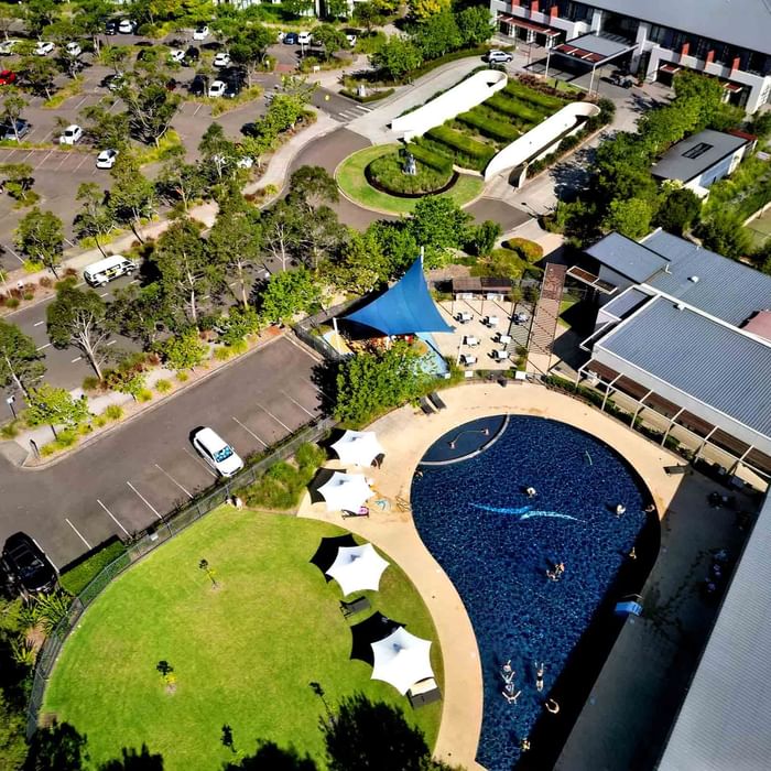 Aerial view of a pool with people swimming, surrounded by loungers, grass, and a parking lot at Mercure Kooindah Waters