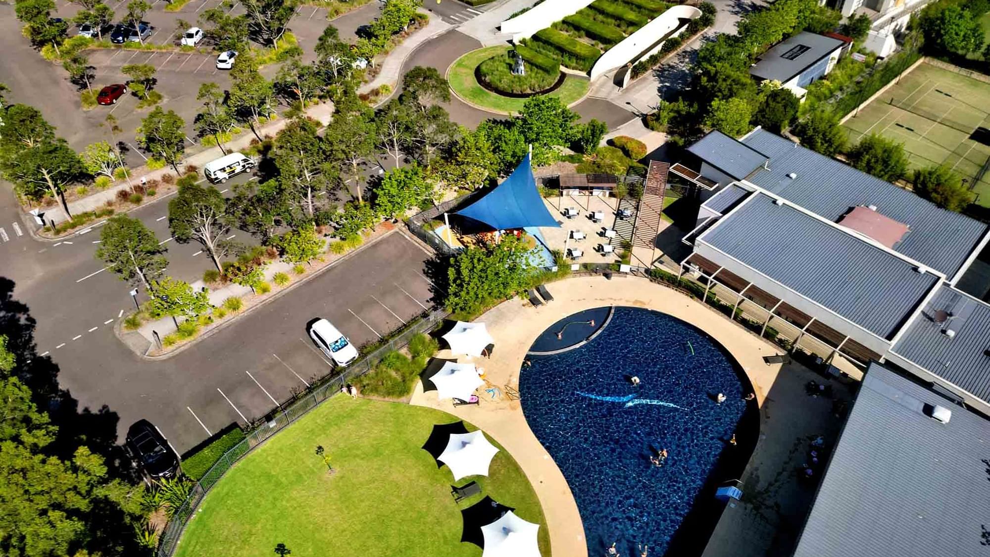 Aerial view of a pool with people swimming, surrounded by loungers, grass, and a parking lot at Mercure Kooindah Waters