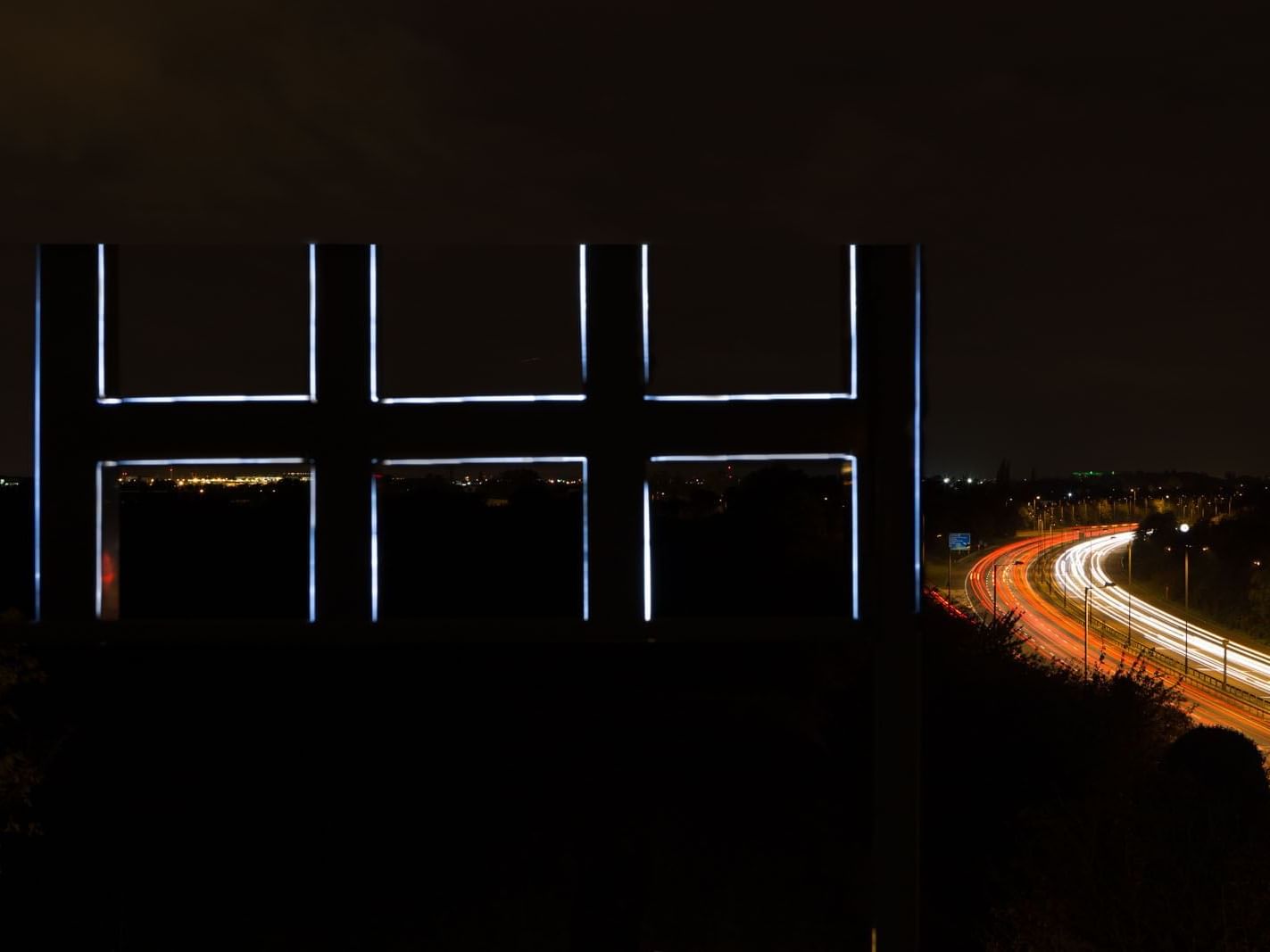 Skyline sculpture with six lit panes overlooks a night highway with glowing car trails and city lights near Heston Hyde Hotel