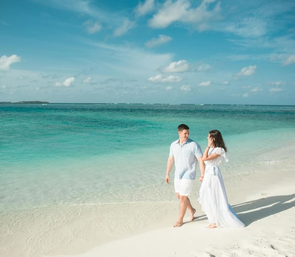 Couple walking hand in hand along a sandy beach with turquoise waters and fluffy clouds at Ananea Madivaru Maldives
