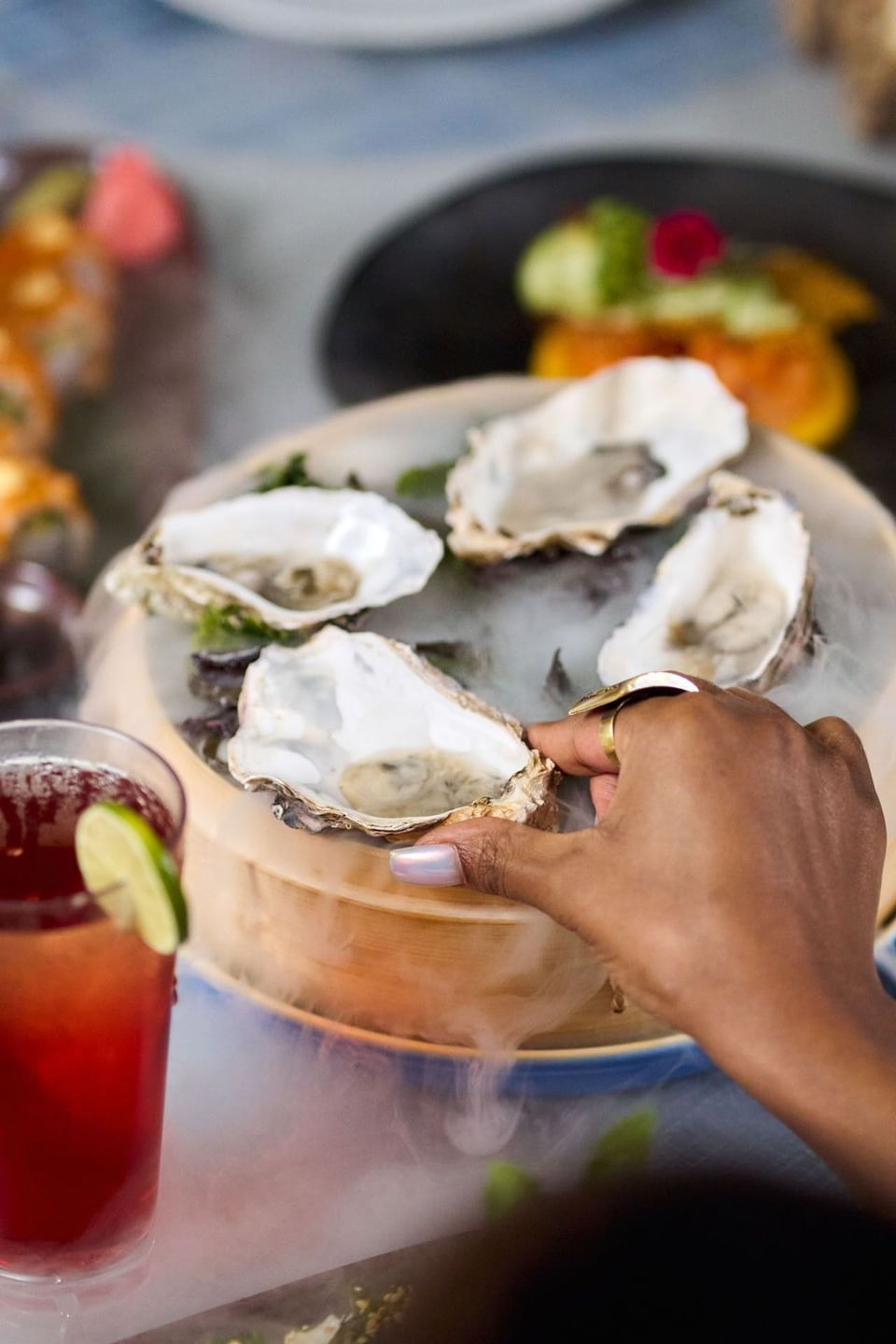 Hand reaching for fresh raw oysters served on a smoking dry ice platter next to an iced cocktail at Malibu Sky Lounge & Bar