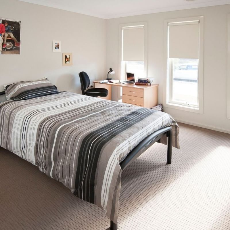 A neat bedroom with a bed, desk, and three windows, featuring a poster on the wall.