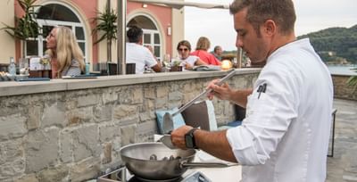 Chef en tenue blanche préparant des aliments sur un gril extérieur avec des clients en arrière-plan.