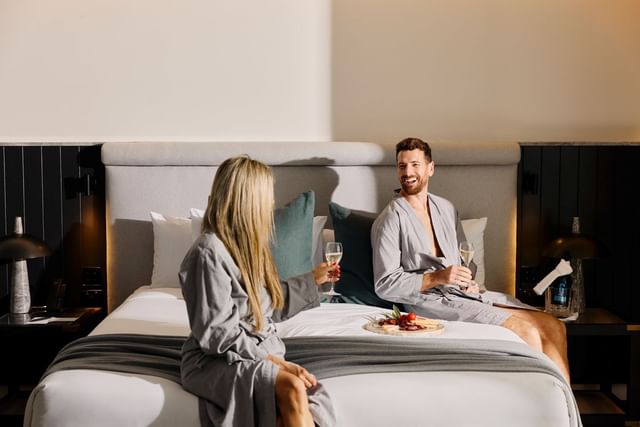 Couple in robes at Elysium Manly drinking wine by a charcuterie platter on a large bed, best places to stay in Manly