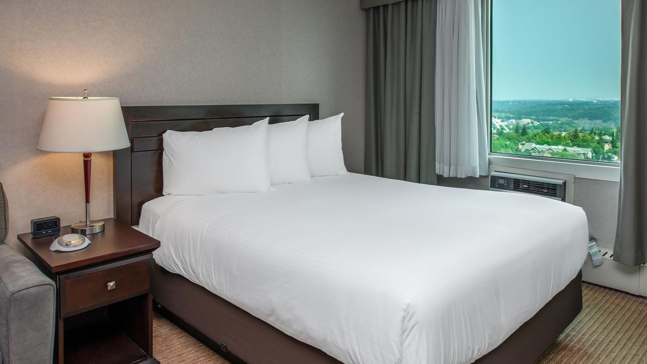 Hotel guest suite with a queen bed, bedside table, lamp and large window with a city view.