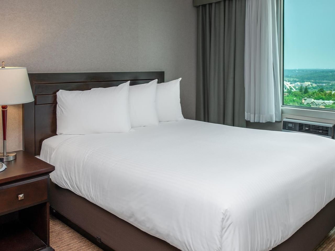 Hotel guest suite with a queen bed, bedside table, lamp and large window with a city view.