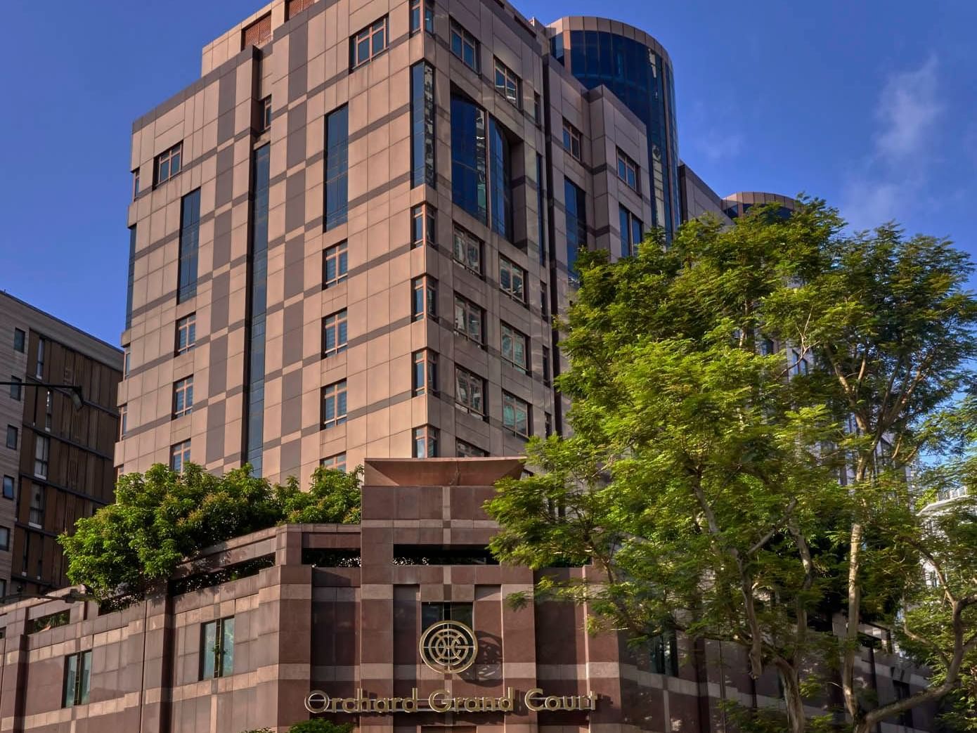 Modern exterior view of Orchard Grand Court, framed by trees under a clear blue sky