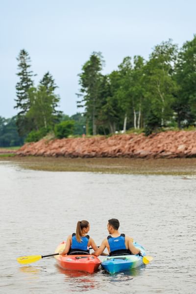 Couple kayaking on calm water by a rocky shoreline under a soft-focus forest at Rodd Brudenell River Resort