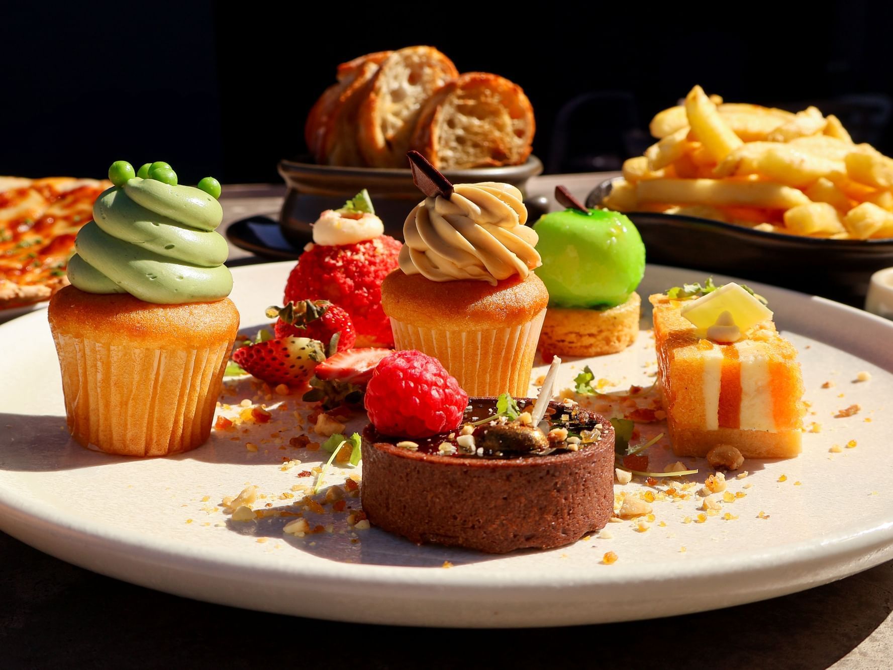 Assorted gourmet desserts on a white plate including cupcakes, chocolate tart, fruit pastries and macarons with pizza and fries in the background.