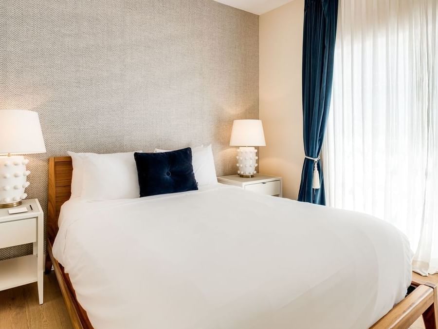 A neatly made bed with white linens and a dark blue pillow in a room with a window and curtains.