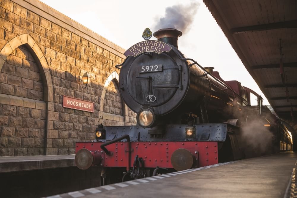 The front of a red and black steam engine labeled "The Hogwarts Express” pulled into a station with a brick wall labeled "Hogsmeade”. 