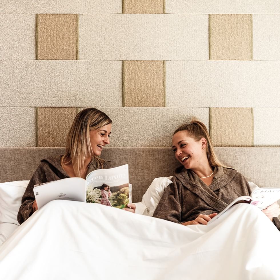 Due donne in accappatoio sul letto con coperte bianche, sorridono leggendo riviste.