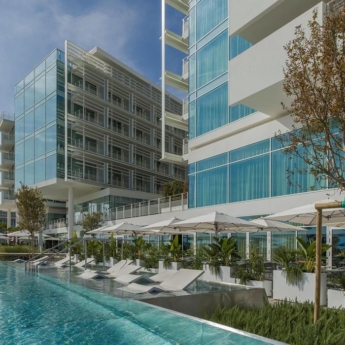 Modern hotel with glass facade, outdoor pool, lounge chairs, umbrellas, and greenery under a blue sky.