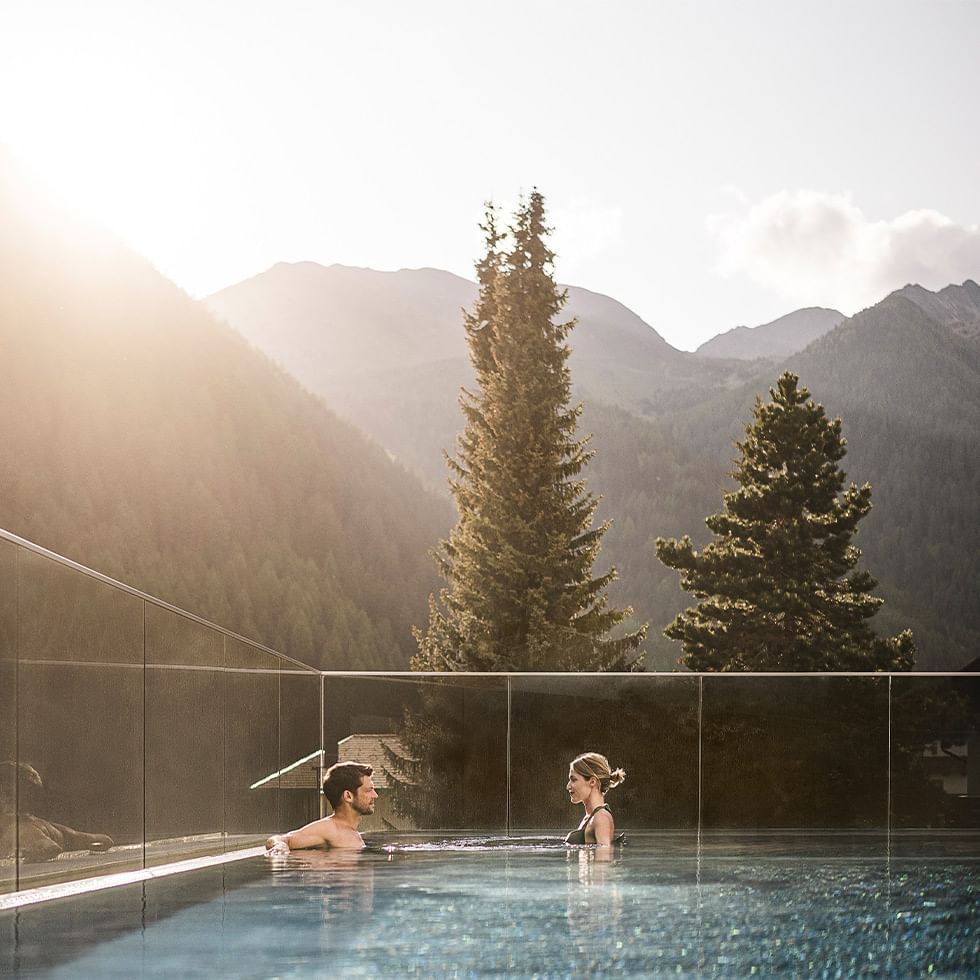 Couple relaxing in outdoor pool with glass walls, surrounded by tall trees and mountains.