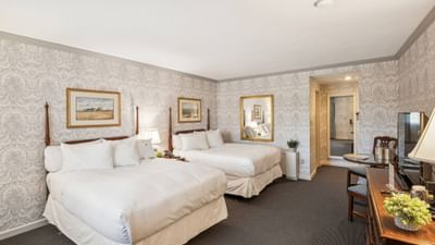 Two double beds against patterned wallpaper near a wooden TV dresser in the guest room at Avon Old Farms Hotel