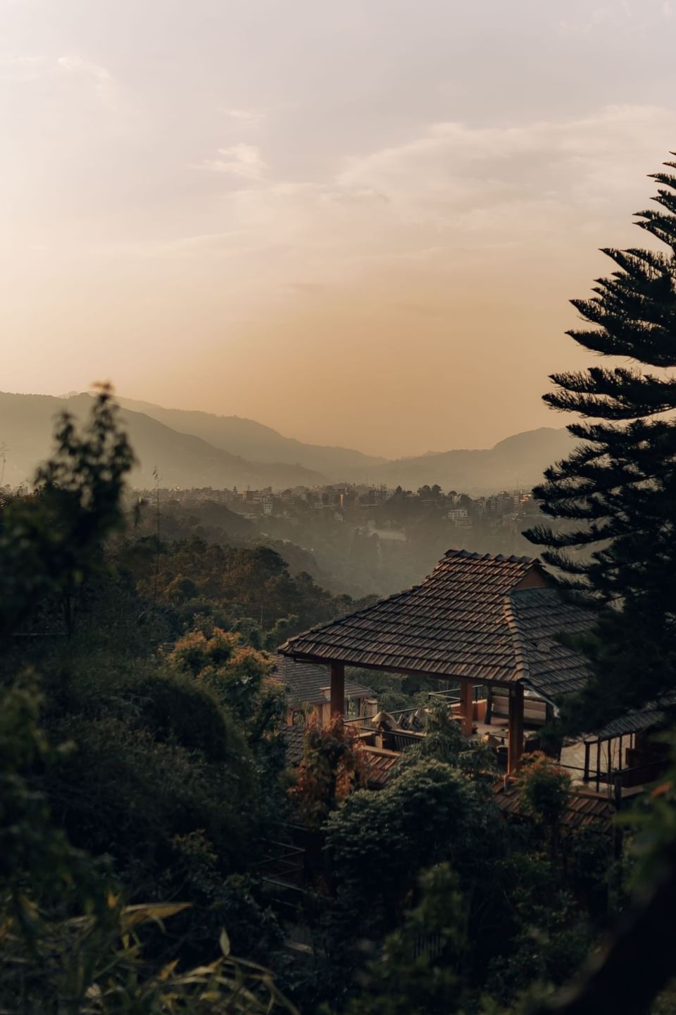 A peaceful hillside view of a wooden house surrounded by lush greenery and mountains near The Dwarika’s Hotels and Resorts