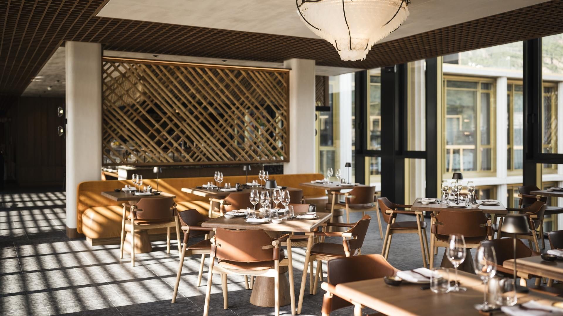 Elegantes Restaurant mit hölzernen Tischen und Stühlen, großen Fenstern und einer modernen Deckenleuchte.