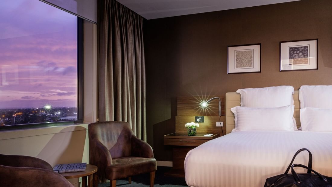 Modern hotel room with twin beds, brown leather chairs, and floor-to-ceiling windows overlooking twilight cityscape