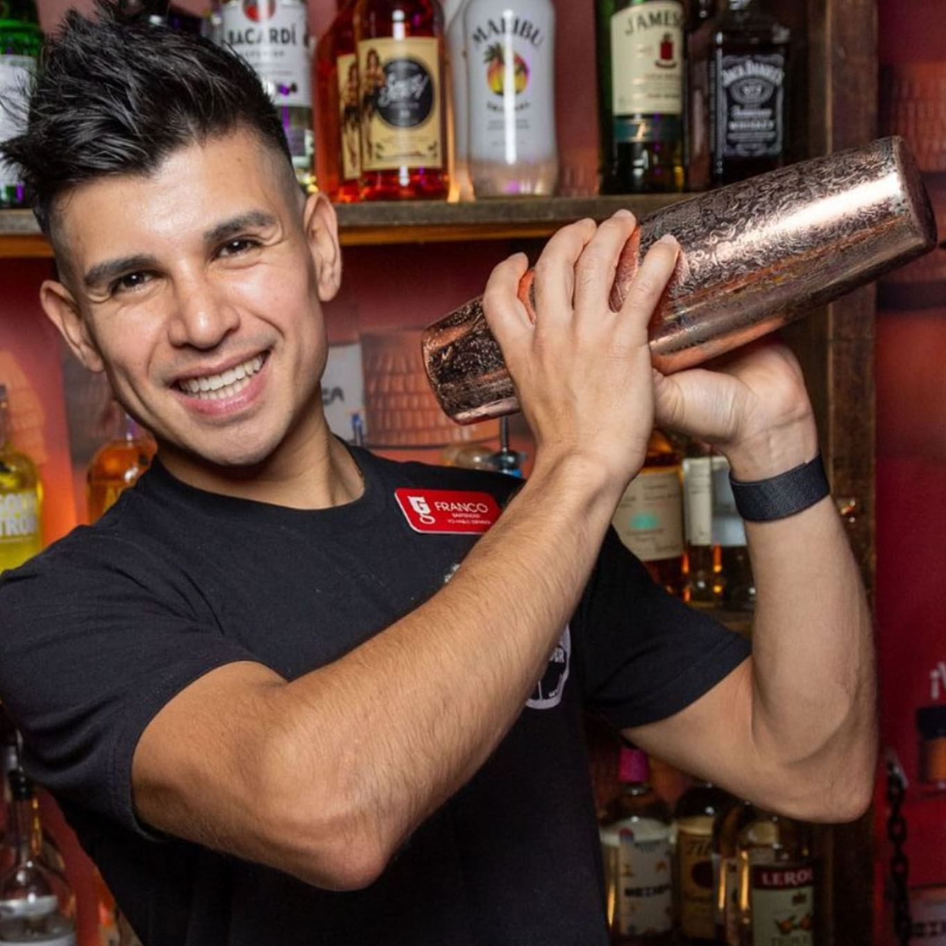 A bartender in a black shirt shakes a cocktail shaker, surrounded by various bottles of alcohol on a shelf at The Gaythering