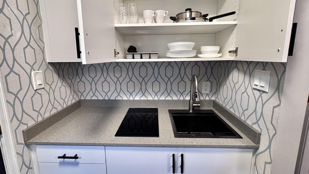 White kitchen with induction cooktop, sink, wall-mounted cabinets, and geometric wallpaper on the wall.