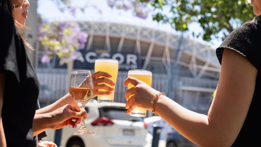 Three people toasting with wine and beer glasses outdoors near a stadium on a sunny day.