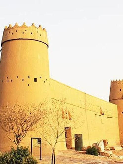 Masmak Citadel with circular towers by a flag and desert plants near Cantonal Hotel by Warwick Riyadh