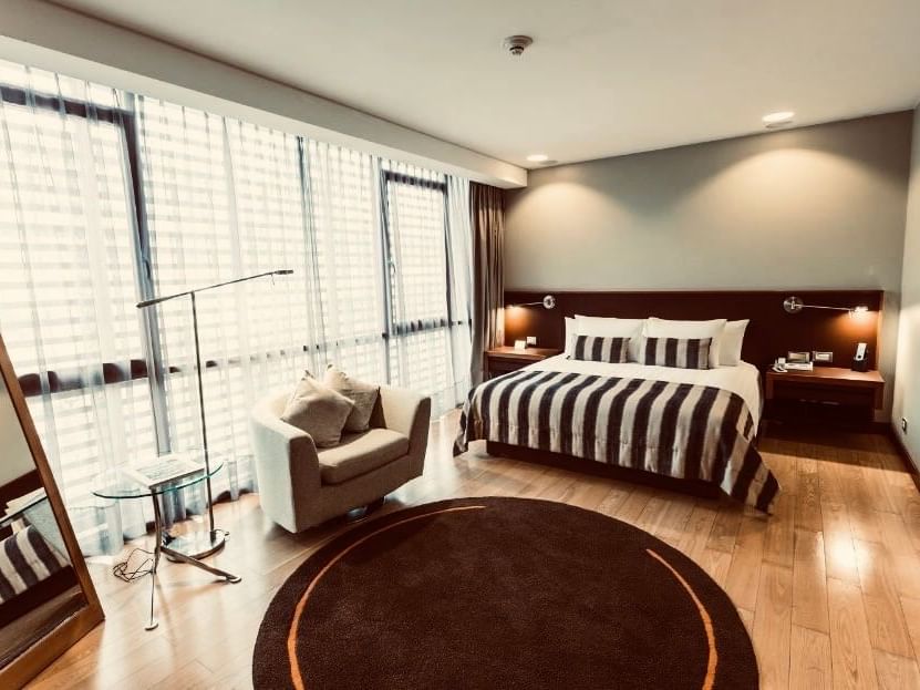 Quarto Deluxe King espaçoso com cama king, poltrona, piso de madeira e grandes janelas no NOI Vitacura em Vitacura